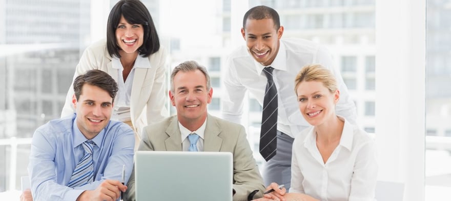 Happy business people gathered around laptop looking at camera in the office